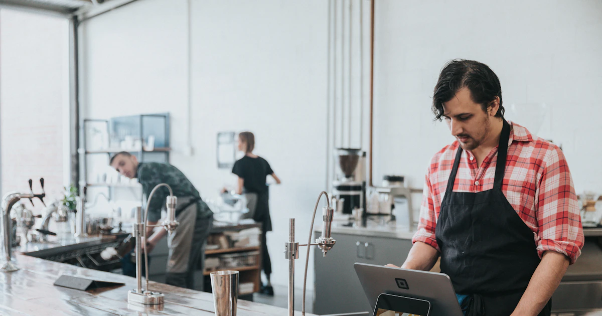 A café worker in a plaid shirt and apron using a POS tablet at a busy service counter