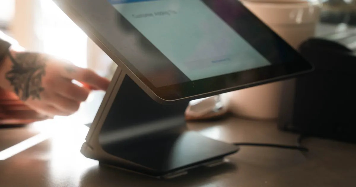 A tablet point-of-sale terminal on a café counter with a staff member's hand reaching toward the screen